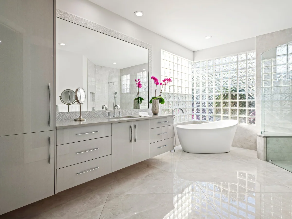Custom primary bath with a full-wall double vanity in satin grey and a freestanding tub at the glass block window